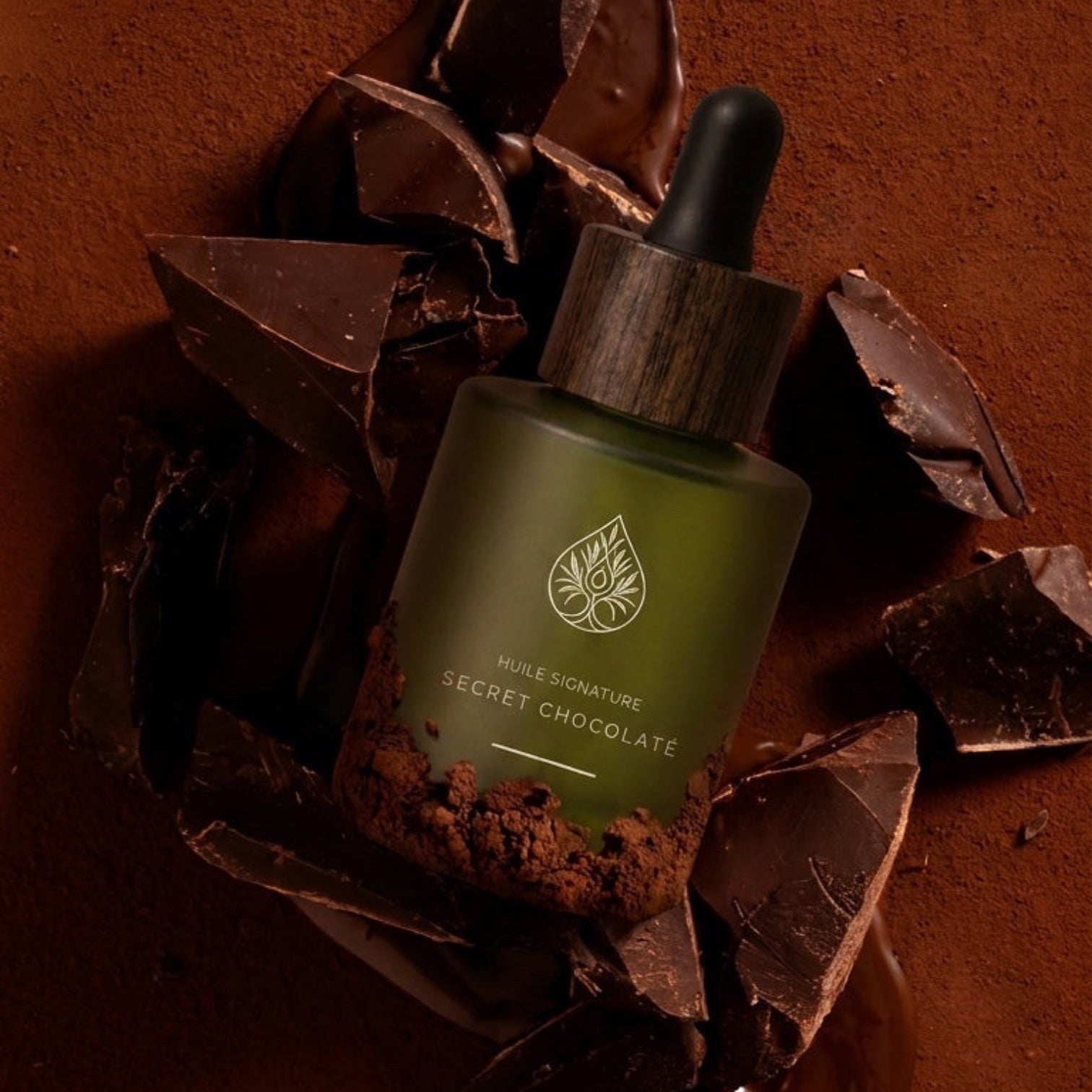 Huile signature Secret Chocolaté L'Arbre de paix dans un flacon en verre dépoli entouré de morceaux de chocolat noir
