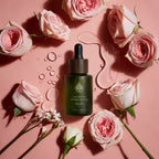 L'Arbre de paix Huile signature Nuage de Rose in a green glass dropper bottle surrounded by fresh pink roses.